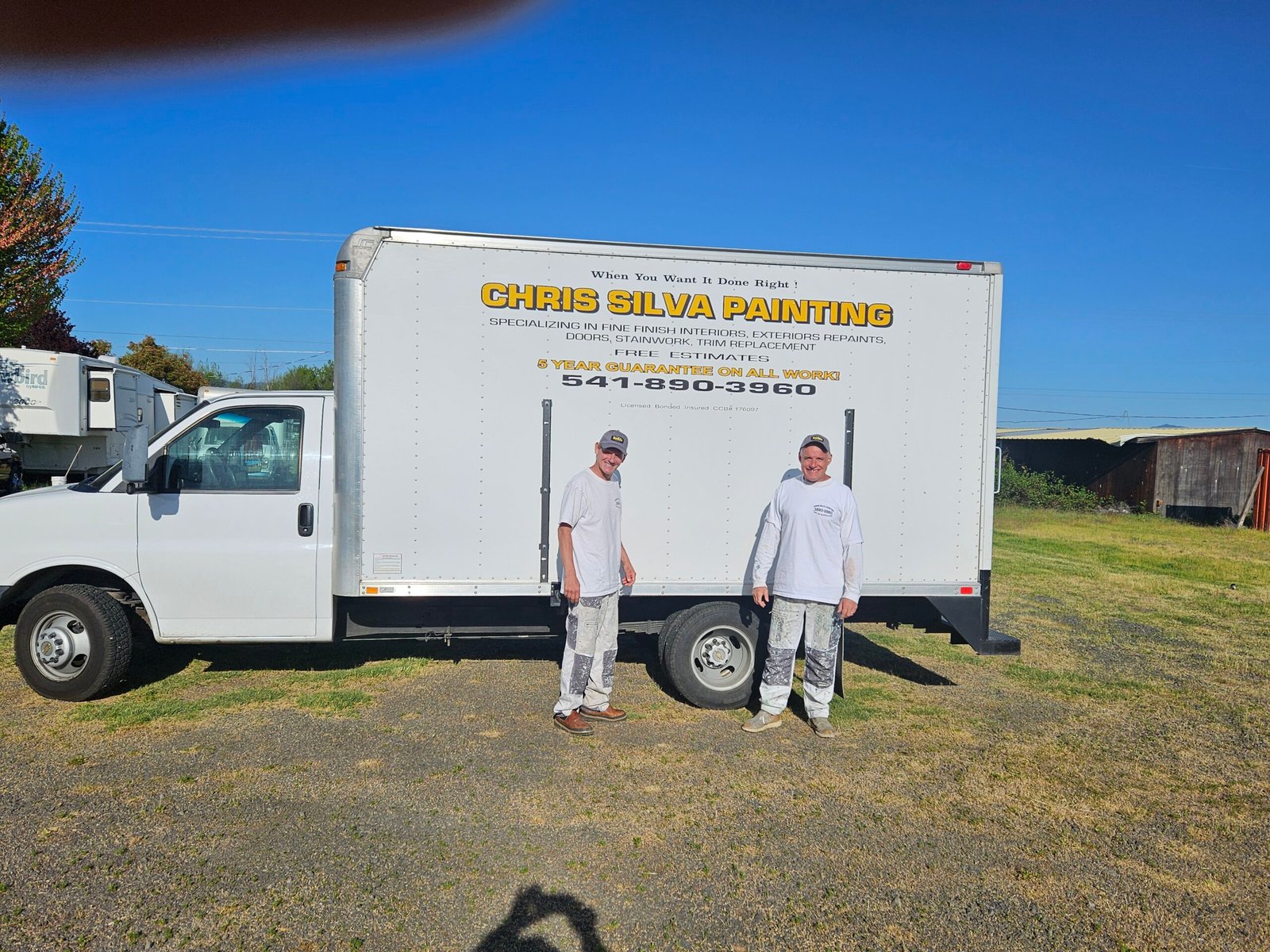 Alt text: Chris Silva Painting box truck parked on a grassy lot in the Rogue Valley, with two painters in white work clothes standing beside the truck displaying the company name and phone number.